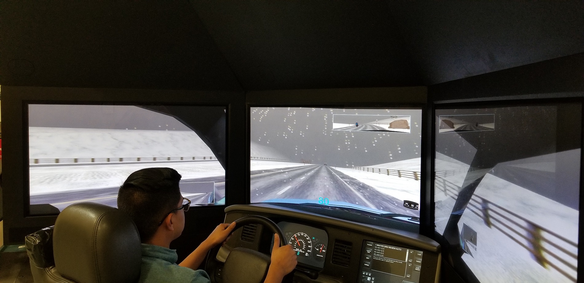 Person using a driving simulator with three large screens displaying a snowy road with guardrails. The dashboard and steering wheel of the simulator are visible, showing a realistic vehicle interior.