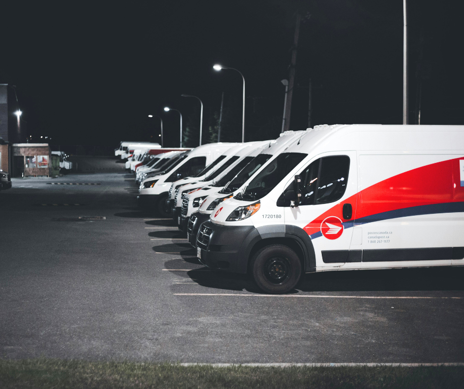 A row of postal delivery vans parked in a lot at night. The vans are white with red and blue markings, aligned neatly under streetlights, creating a uniform appearance against the dark background. A row of postal delivery vans parked in a lot at night. The vans are white with red and blue markings, aligned neatly under streetlights, creating a uniform appearance against the dark background.