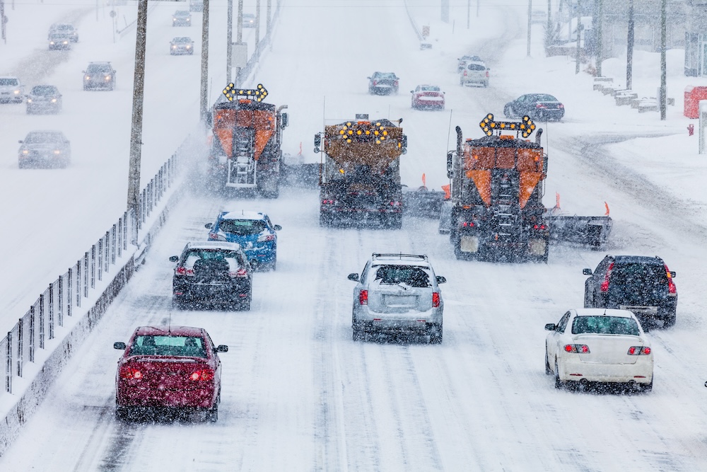 Cars follow two snow plows clearing a multi-lane road during heavy snowfall. The road and surroundings are covered in snow, and visibility is reduced. Traffic moves cautiously behind the plows in low light conditions.