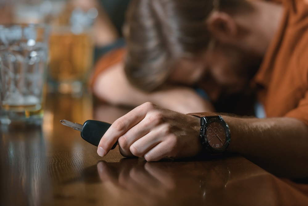 A man with his head down on a bar counter holds car keys. Glasses of beer are visible nearby.