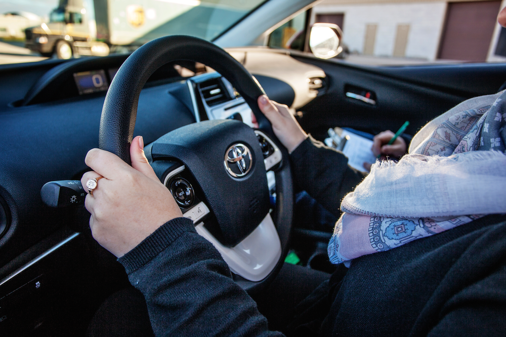 A person wearing a scarf is sitting in the driver's seat of a car, holding the steering wheel with both hands. A pen and notepad are visible in their lap. The car's dashboard, including the Toyota logo, is also visible.