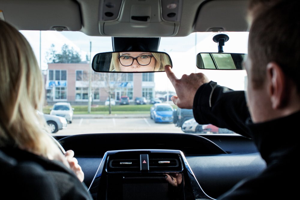 A person in the driver's seat adjusts the rearview mirror, focusing on a woman with glasses reflected in it. The car interior and dashboard are visible, with a parking lot and buildings outside the window.