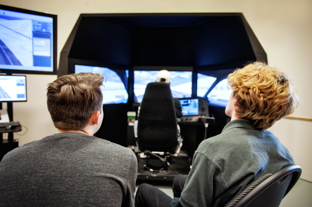 Two people observe a third person seated inside a flight simulator. Multiple screens surround the seated person, displaying simulated flight visuals. Additional monitors are visible to the left, showing detailed interface information.