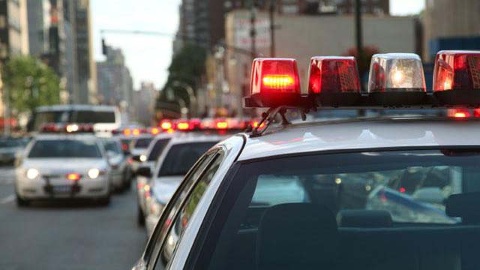 Police cars with flashing lights are in traffic on a city street. The scene is set in an urban area with buildings and other vehicles in the background.