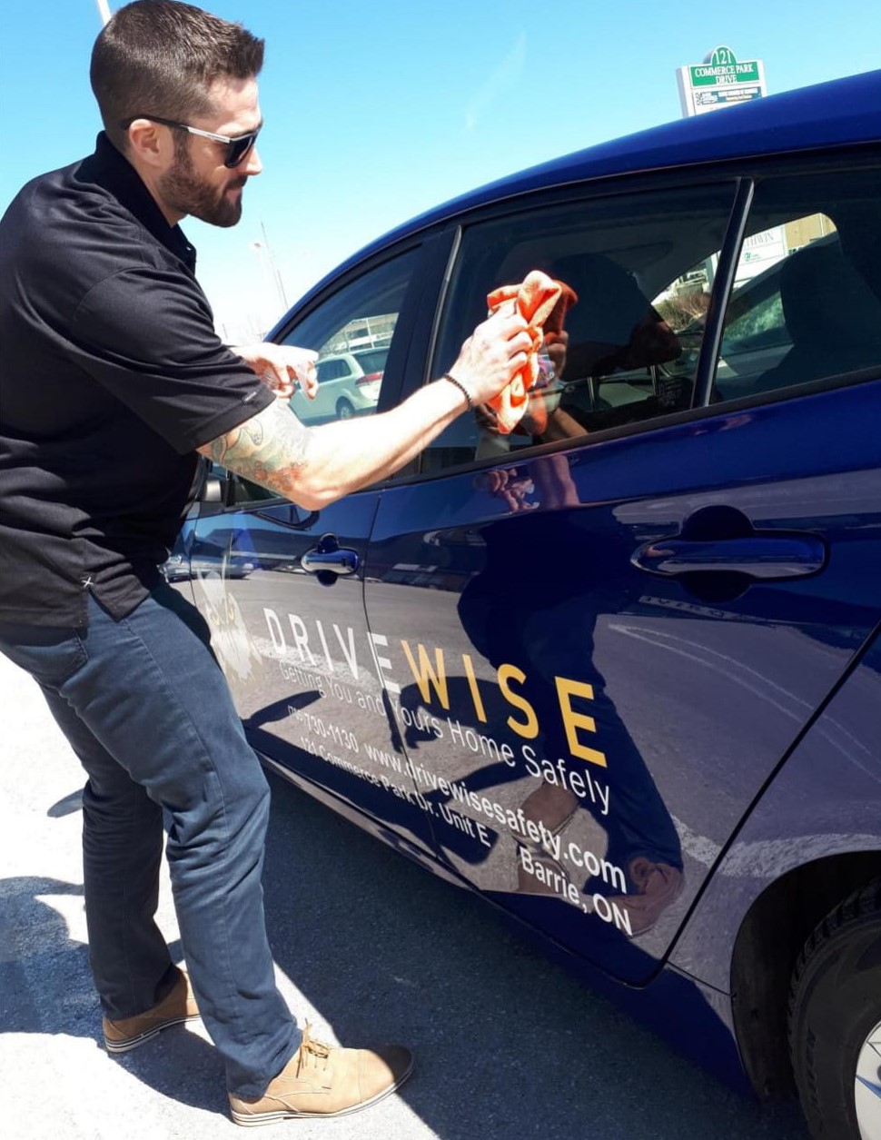 A man wearing sunglasses and a black shirt cleans the window of a blue car with an orange cloth. The car has “Drive Wise” and other text on the side. The scene takes place outdoors on a sunny day. A man wearing sunglasses and a black shirt cleans the window of a blue car with an orange cloth. The car has "Drive Wise" and other text on the side. The scene takes place outdoors on a sunny day.
