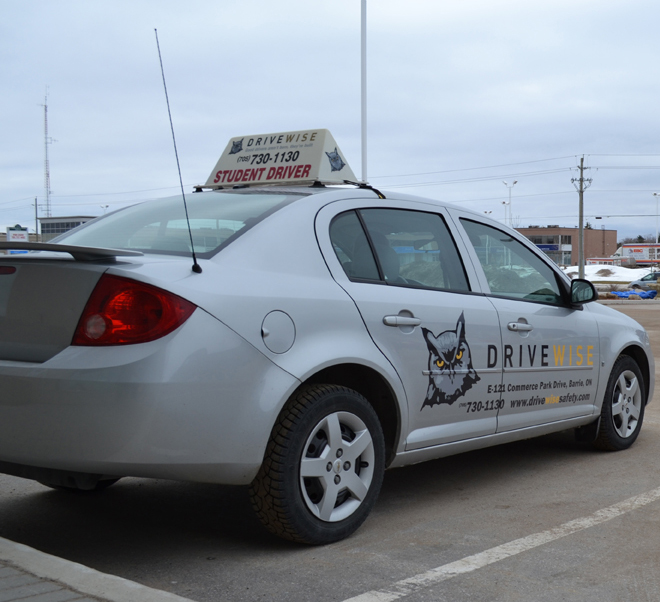 A silver car parked in a lot with a "Student Driver" sign on the roof. The car has the logo of an owl and the brand "DRIVEWISE" on the side. Buildings and a cloudy sky are in the background.