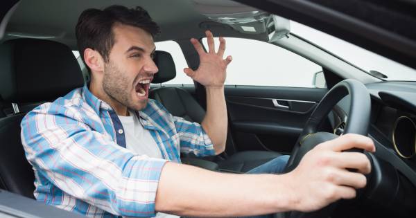 A man in a plaid shirt sits in a car, gripping the steering wheel with one hand and raising the other, looking frustrated or angry. His mouth is open as if shouting, and his expression shows agitation. A man in a plaid shirt sits in a car, gripping the steering wheel with one hand and raising the other, looking frustrated or angry. His mouth is open as if shouting, and his expression shows agitation.