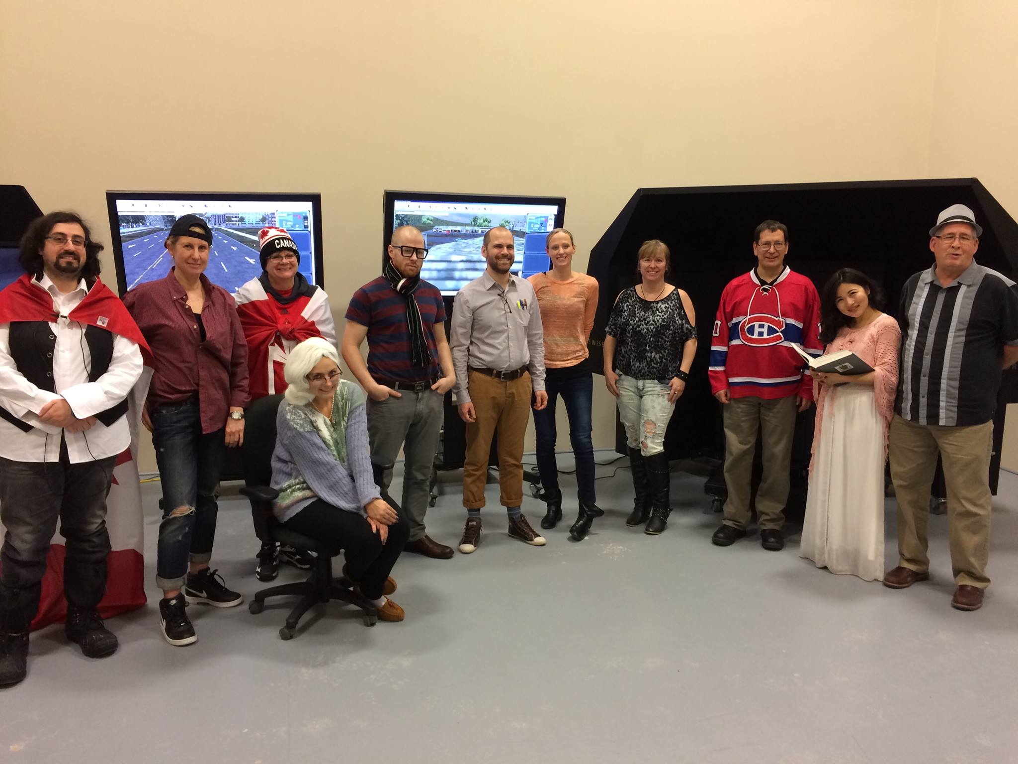 A group of eleven people is posing in a room with three screens behind them. Some are wearing costumes, including a superhero cape, a hockey jersey, and a formal dress. They are standing on a gray floor in front of beige walls.