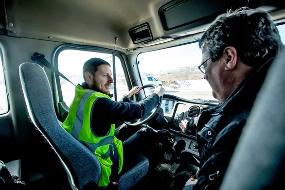 A man wearing a safety vest sits in the driver's seat of a truck, speaking to another man seated beside him who wears glasses. They appear to be engaged in a discussion, with daylight visible through the windshield.