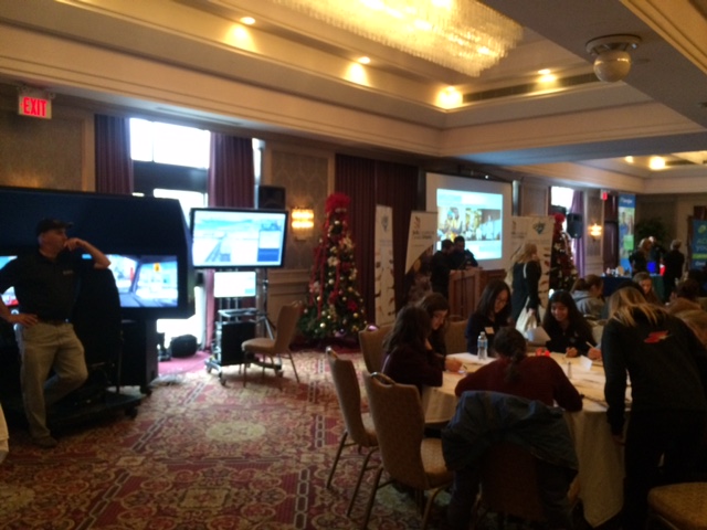 A conference room with people seated at tables, engaging in discussions. Presentation screens and festive Christmas trees are visible. A person stands near a booth with large monitors. The room is well-lit with chandeliers. A conference room with people seated at tables, engaging in discussions. Presentation screens and festive Christmas trees are visible. A person stands near a booth with large monitors. The room is well-lit with chandeliers.