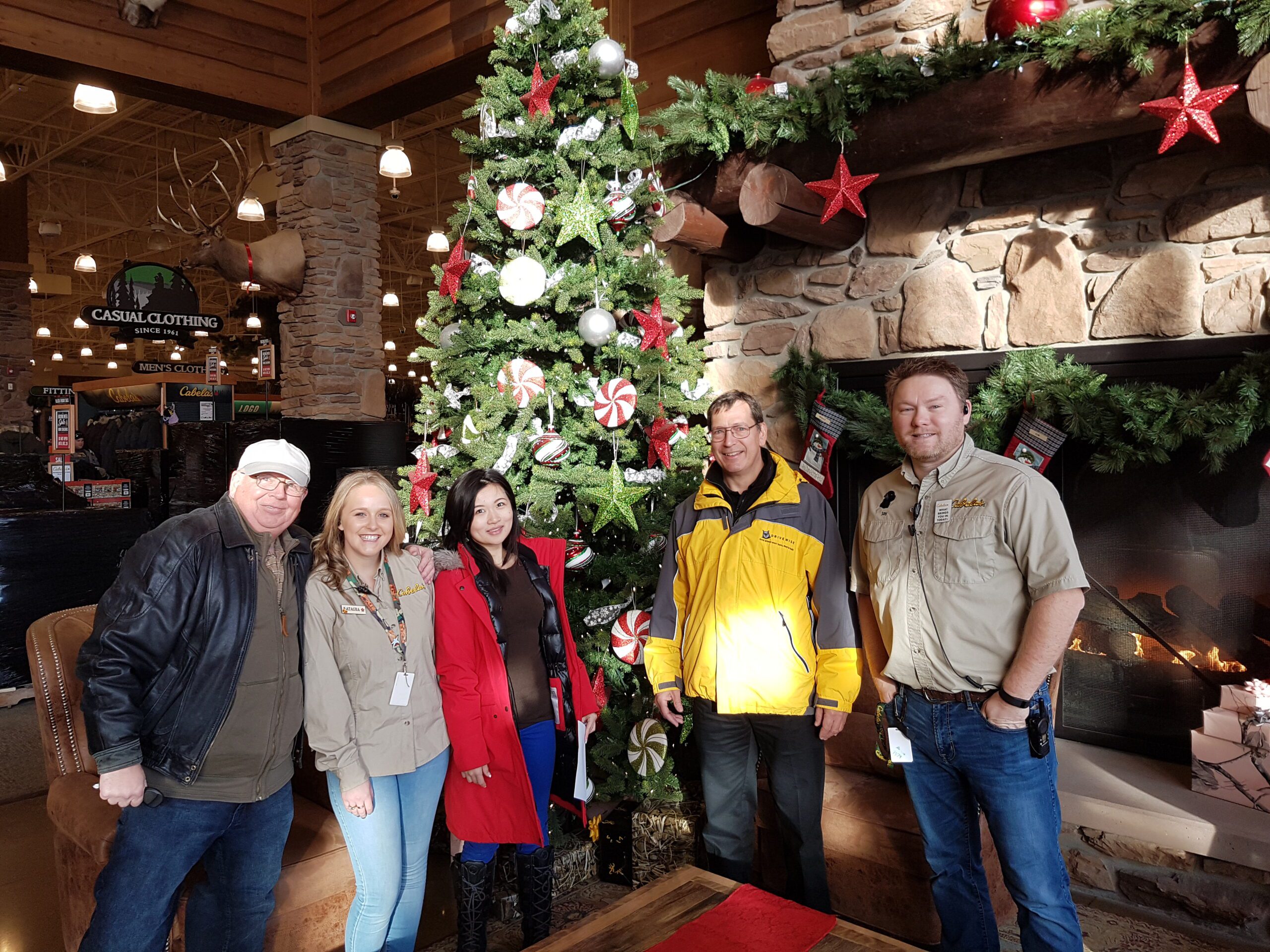 Five people stand in front of a decorated Christmas tree with red and white ornaments inside a rustic store. They are smiling and wearing casual clothes. A stone fireplace and holiday decorations are in the background. Five people stand in front of a decorated Christmas tree with red and white ornaments inside a rustic store. They are smiling and wearing casual clothes. A stone fireplace and holiday decorations are in the background.