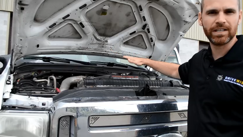A person stands next to a truck with the hood open, pointing at the engine. The truck's front grille and engine components are visible. The person is wearing a black shirt.