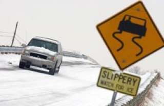 A vehicle drives carefully on a snow-covered road. A yellow warning sign in the foreground shows a car with skid marks and reads "Slippery, Watch for Ice." The scene is overcast, indicating icy conditions.