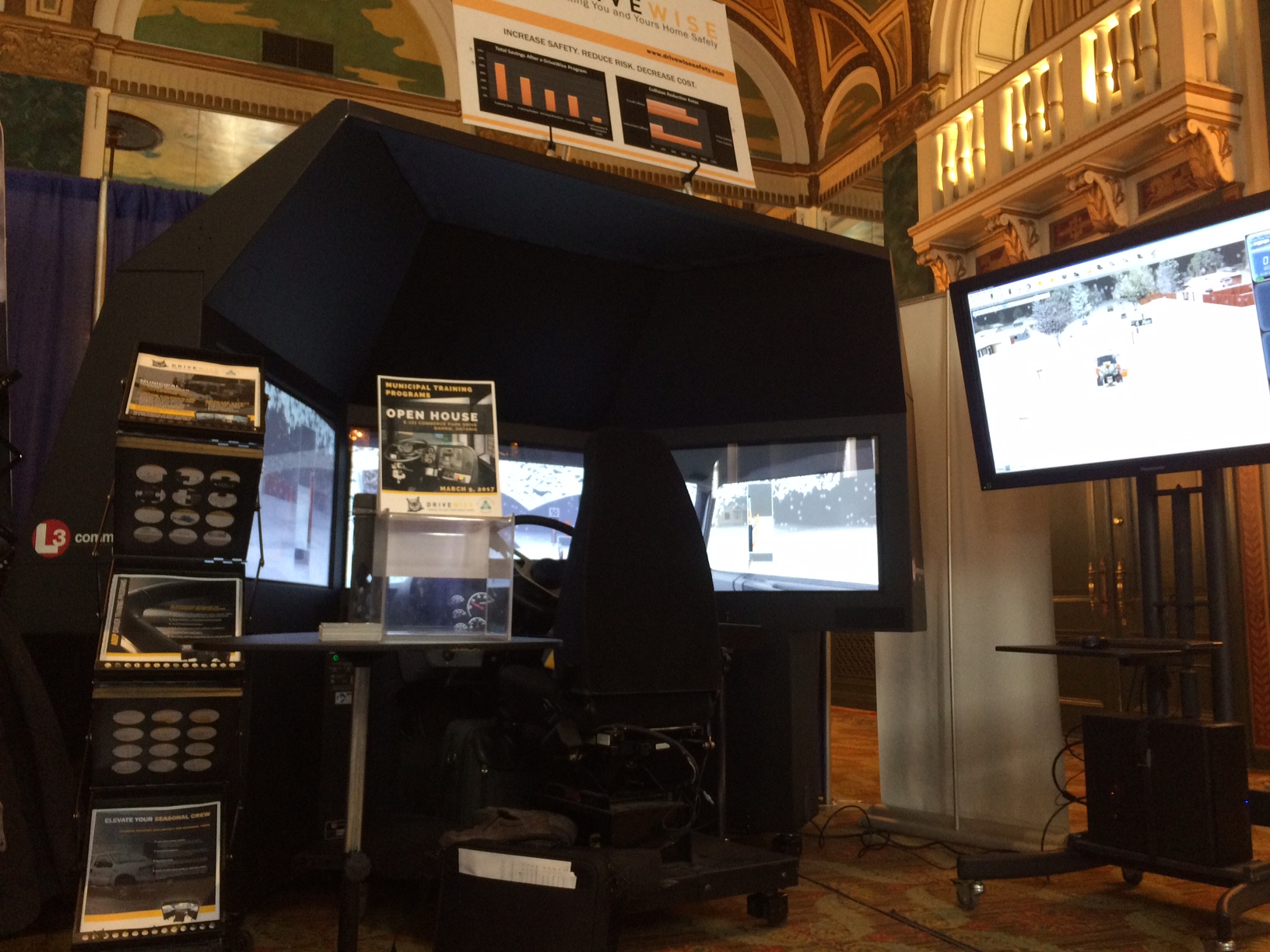 A driving simulator setup is displayed at a booth. It includes multiple screens showing a snowy road scene, and a steering wheel. A large monitor on the right mirrors the simulator’s display. Banners and informational posters are visible around the setup. A driving simulator setup is displayed at a booth. It includes multiple screens showing a snowy road scene, and a steering wheel. A large monitor on the right mirrors the simulator's display. Banners and informational posters are visible around the setup.