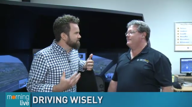 Two men stand in conversation inside a room with computer monitors. One wears a checkered shirt and gestures with his hands, while the other wears a black polo shirt. A sign with driving tips is visible in the background. Text reads “DRIVING WISELY. Two men stand in conversation inside a room with computer monitors. One wears a checkered shirt and gestures with his hands, while the other wears a black polo shirt. A sign with driving tips is visible in the background. Text reads "DRIVING WISELY.