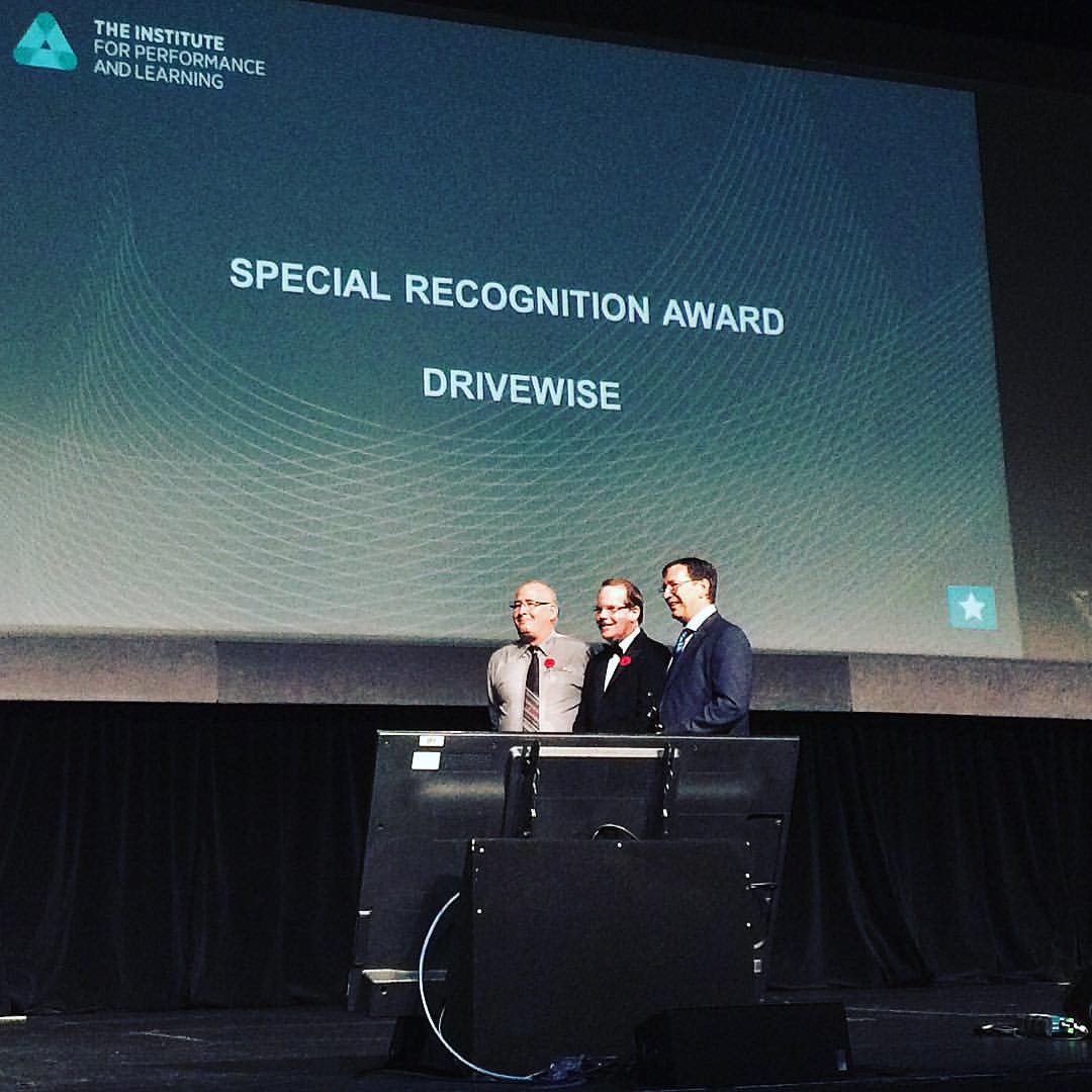 Three men stand on a stage in front of a large screen displaying "Special Recognition Award Drivewise" at the Institute for Performance and Learning event. A podium is in front of them.