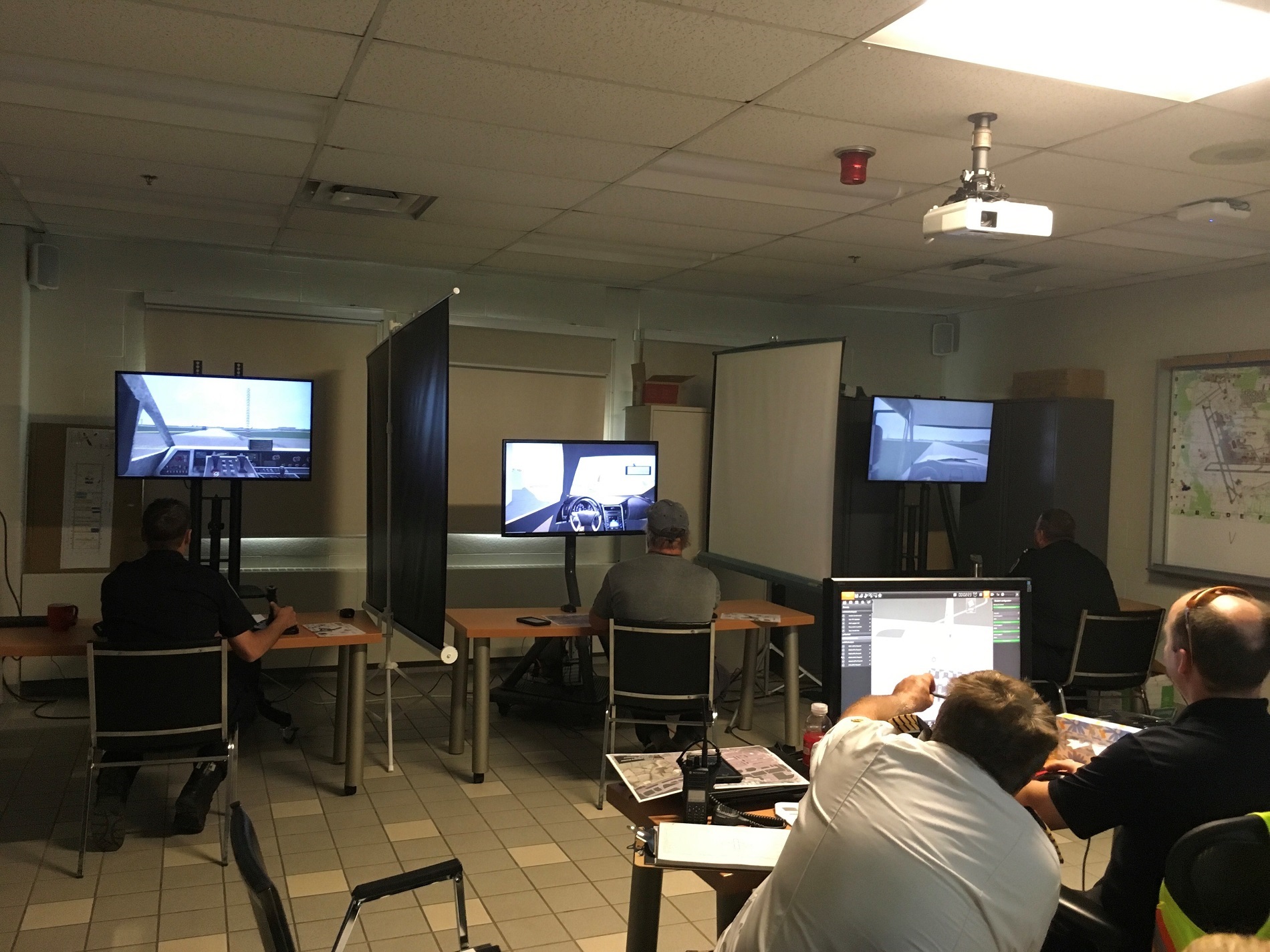 A room with four people seated at desks, each using a computer with monitors displaying vehicle simulation software. The room has a ceiling projector and maps on the wall. Dividers separate some of the workstations.