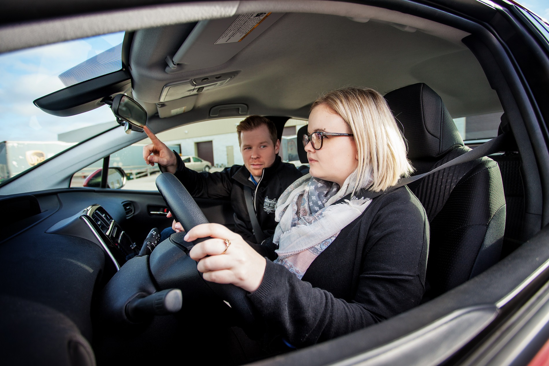 A woman sits in the driver's seat of a car, hands on the steering wheel. A man in the passenger seat points at the rearview mirror, appearing to give instructions. Both are focused, suggesting a driving lesson.
