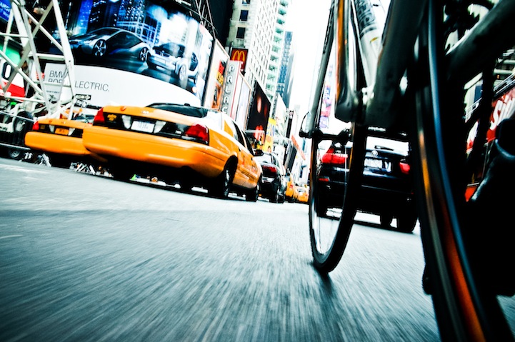 Close-up view of a bicycle wheel in motion on a busy city street, with yellow taxis and other vehicles. Skyscrapers and bright billboards, including a McDonald's sign, fill the background, suggesting an urban atmosphere.