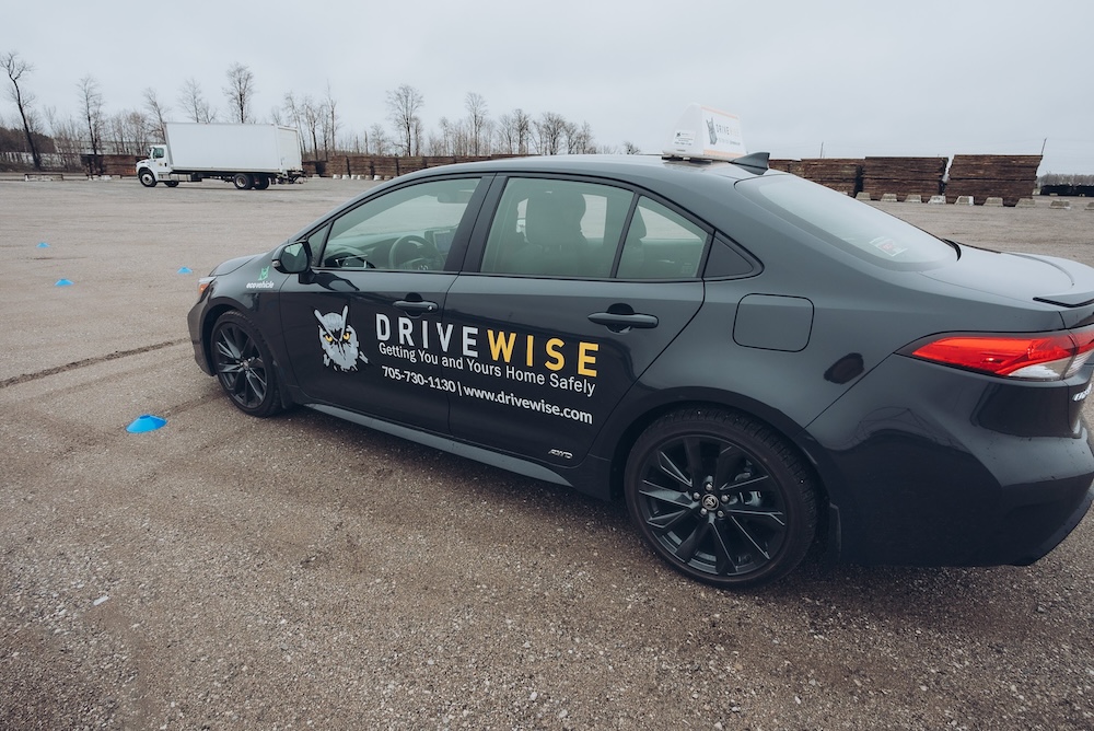 A gray sedan with the DRVIEWISE logo and contact details on its side is parked on an empty lot. Blue cones are scattered around, suggesting a driving course. Trees and a fence are visible in the background under an overcast sky.