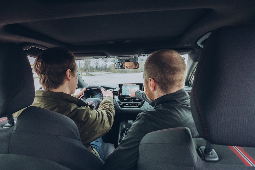 Two people are sitting in the front seats of a car. The person on the left is driving and pointing at the dashboard, while the person on the right is also pointing at the screen in the center console. The car interior is visible.