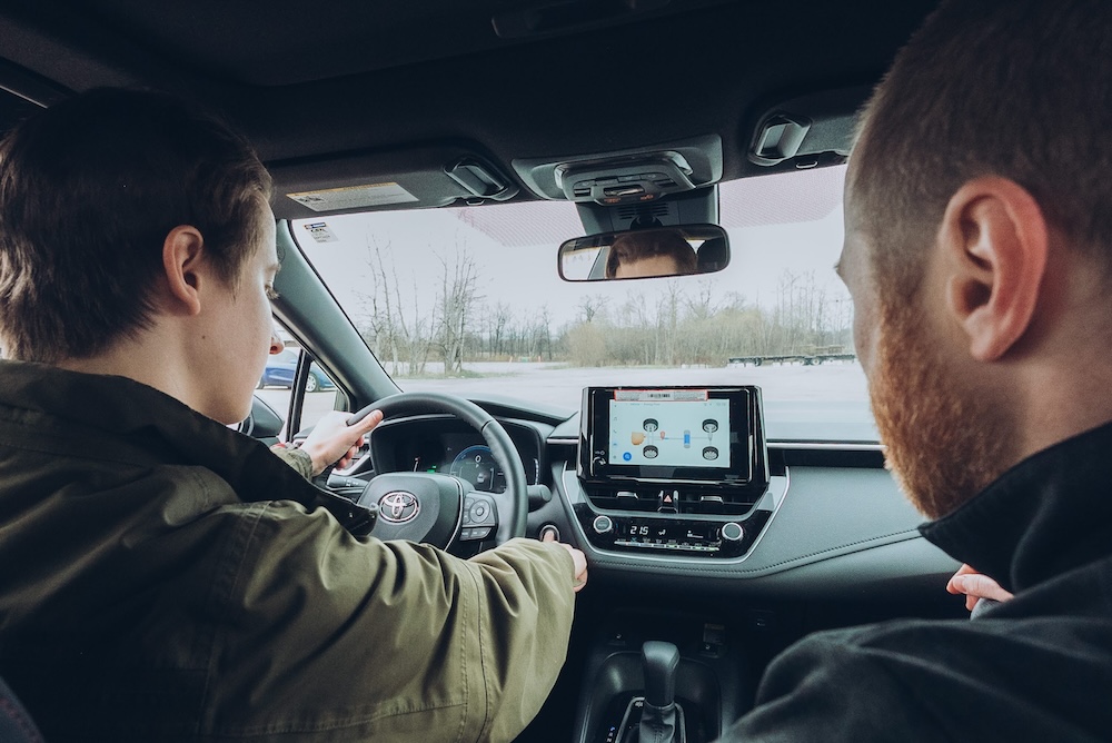 Two people sit in the front seats of a car. One is driving while the other observes. The dashboard features a touchscreen displaying a map. Trees are visible through the windshield.