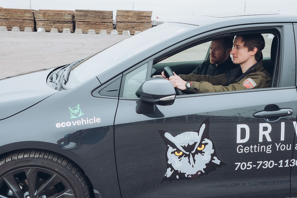 Two people sit in a parked car labeled "ecovehicle" and "Driver" with an owl logo. The driver is a young person wearing a green jacket, and the passenger is an older person wearing a black jacket. They are in an open, paved area.
