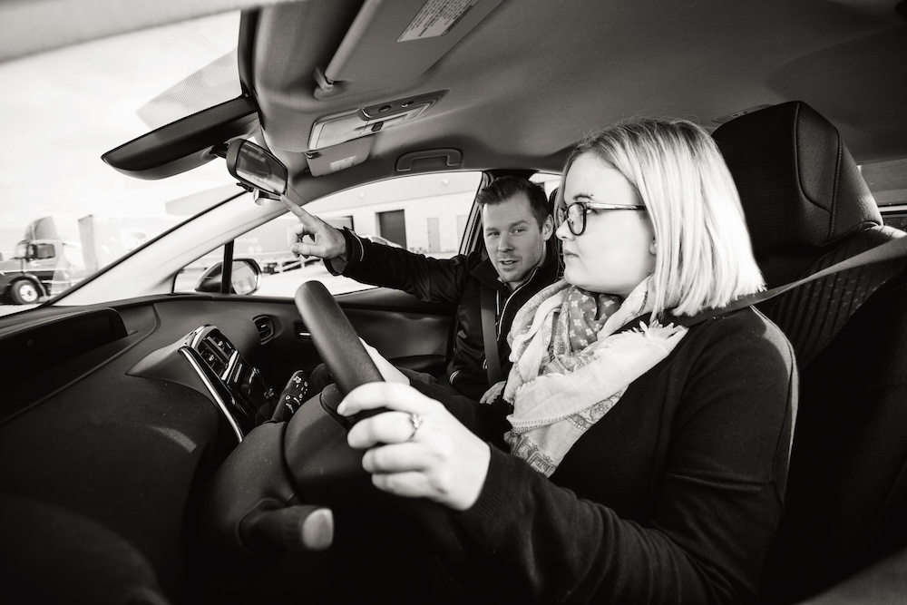 A woman is sitting in the driver's seat of a car, holding the steering wheel, while a man in the passenger seat points at the rearview mirror. They appear to be discussing something inside the car.