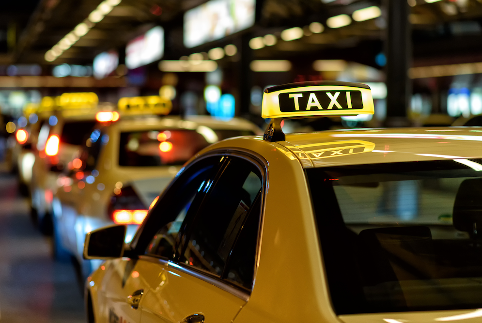 A line of yellow taxis with illuminated taxi signs waits in a dimly lit urban setting. The focus is on the nearest taxi's sign, while blurred lights in the background suggest a bustling city atmosphere.
