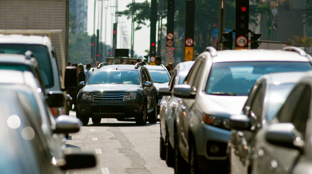 A busy urban street with traffic congestion shows a variety of cars stuck in a jam. A dark SUV is prominently visible in the center. Pedestrians are seen on the sidewalk, and trees are visible in the background with traffic lights overhead.