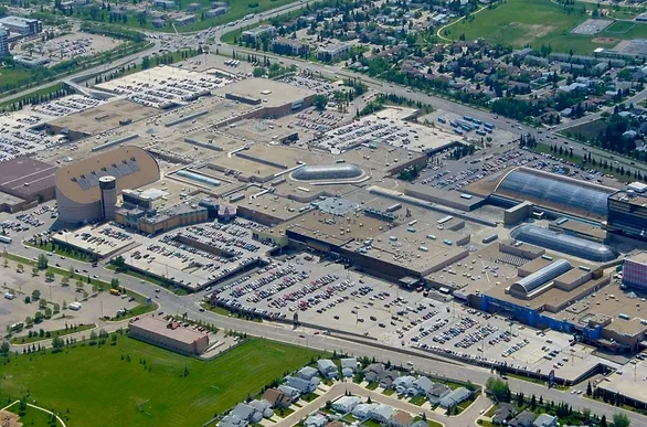 Aerial view of a large shopping mall with expansive parking lots and surrounding green areas. The building features unique dome and curved structures. Nearby residential areas and roads are visible, creating a bustling urban scene.
