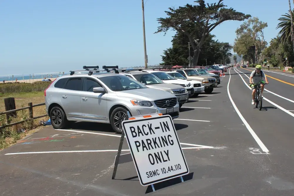A row of cars parked diagonally along a road next to a beach is marked with a "Back-In Parking Only" sign in the foreground. A cyclist wearing a helmet rides on a nearby bike lane. Palm trees and ocean water are visible in the background.