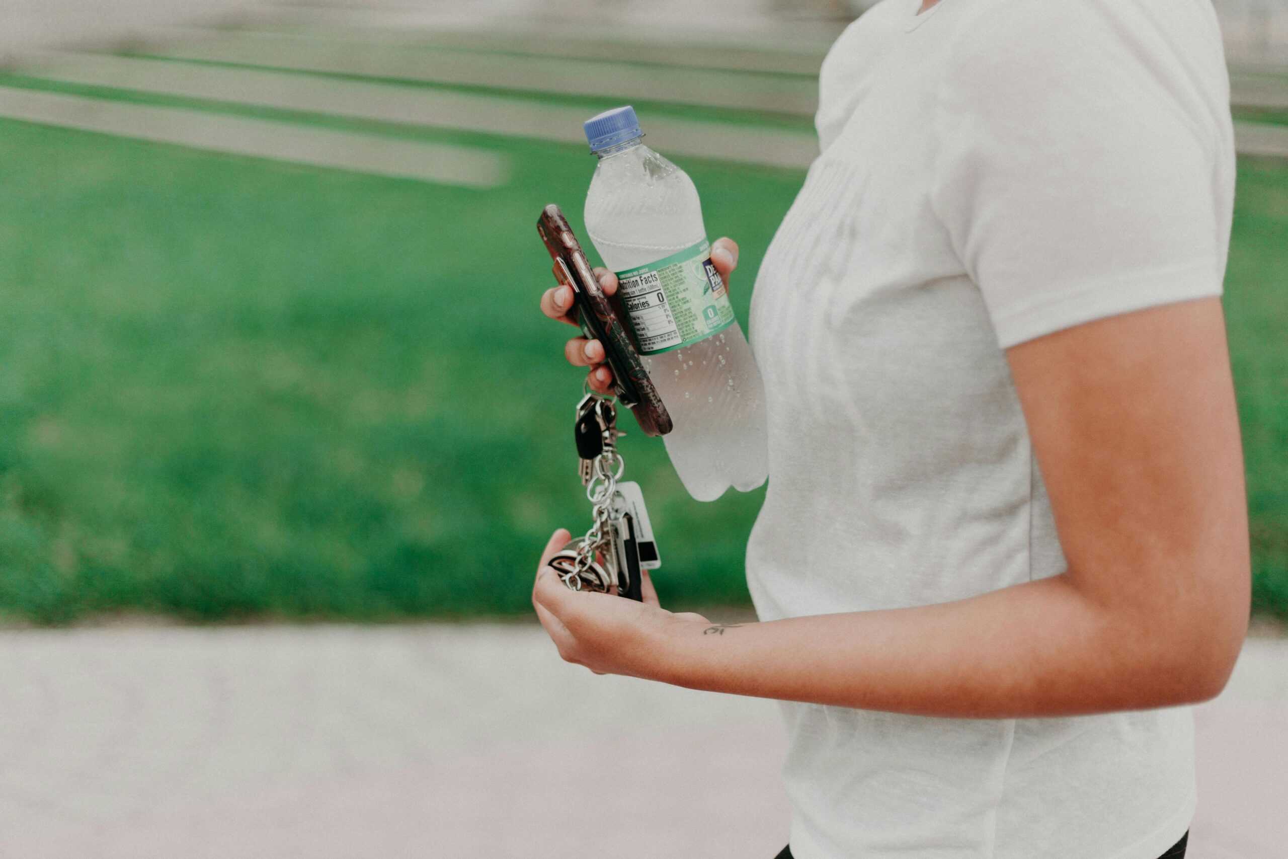 A person in a white shirt holds a smartphone with a patterned case, a plastic water bottle, and a keychain in one hand. They are walking on a path with green grass and stone tiles in the background.