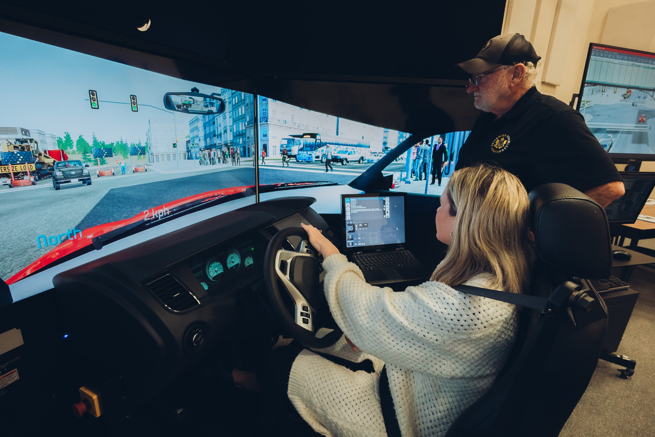 A woman sits at a driving simulator with a steering wheel, while a man stands beside her, observing. Multiple screens display a virtual city street, and a laptop is set up nearby for monitoring.