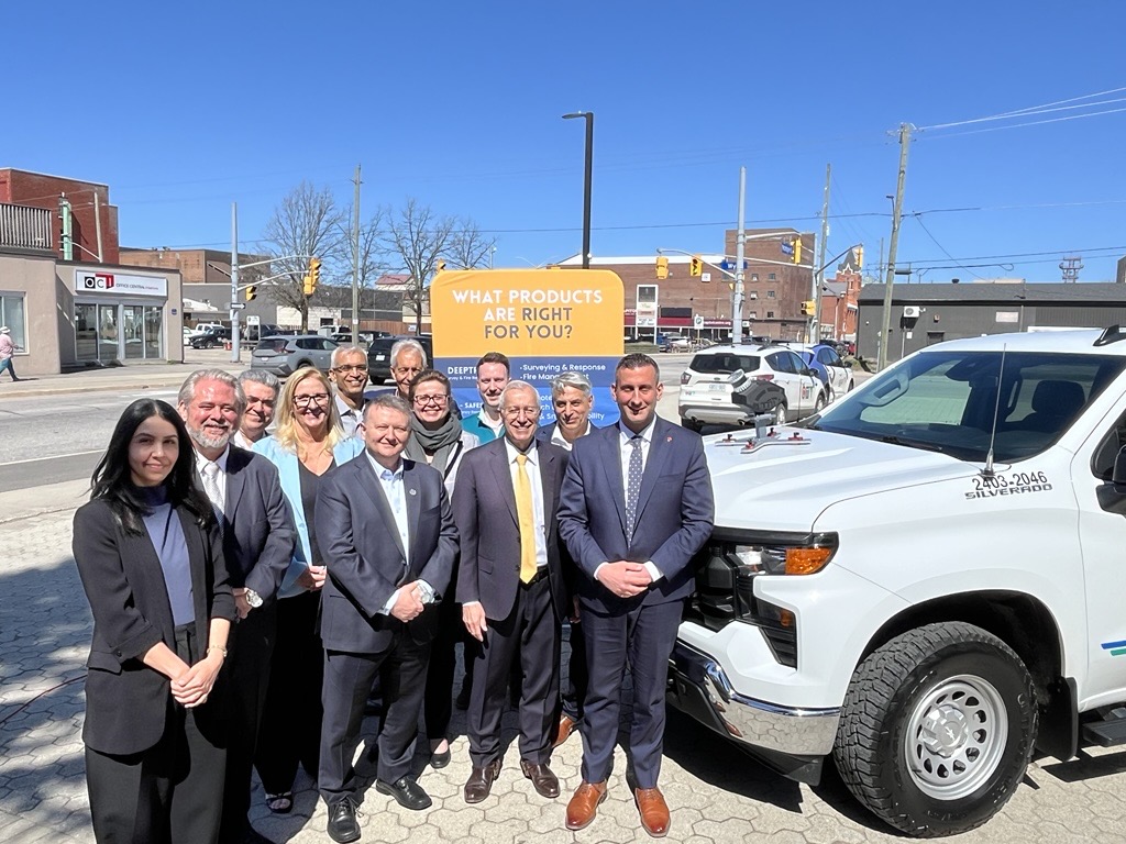 A group of people in business attire stand smiling on a sidewalk next to a white pickup truck under a clear blue sky, with shops and an orange sign reading “WHAT PRODUCTS ARE RIGHT FOR YOU?”—promoting OVIN and VR snowplow training—in the background.