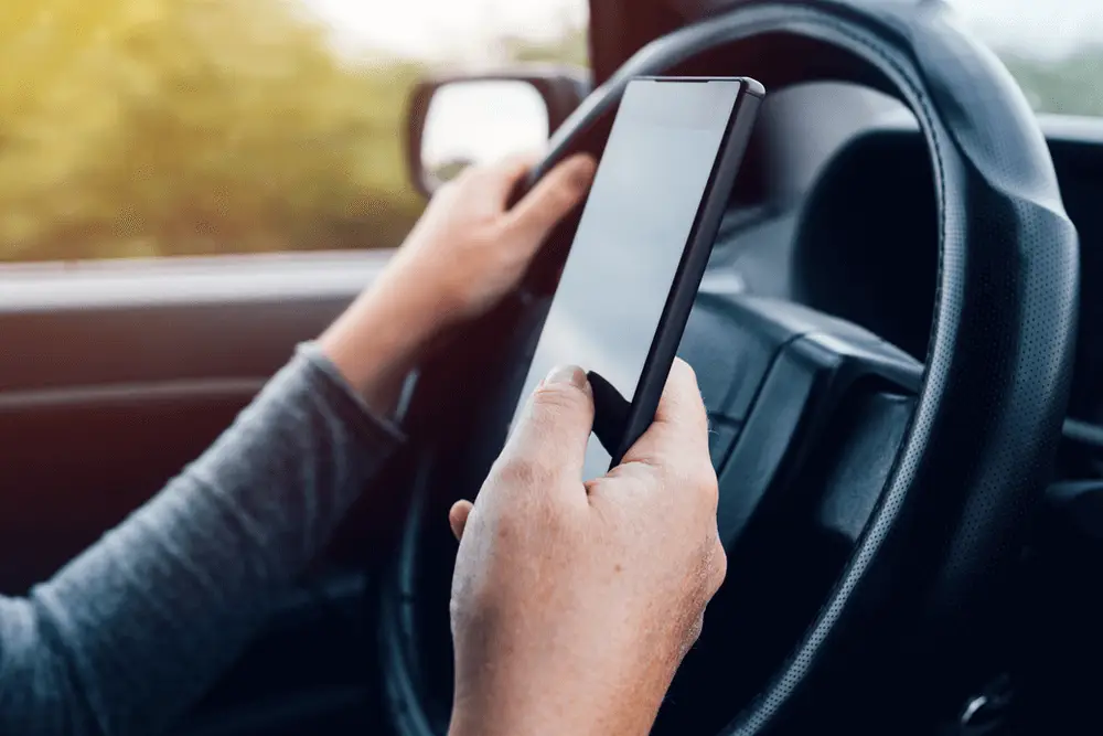Closeup-driver-holding-a-phone-DriveWise A person holds a smartphone in one hand while driving, with the other hand on the steering wheel, suggesting distracted driving inside a car.