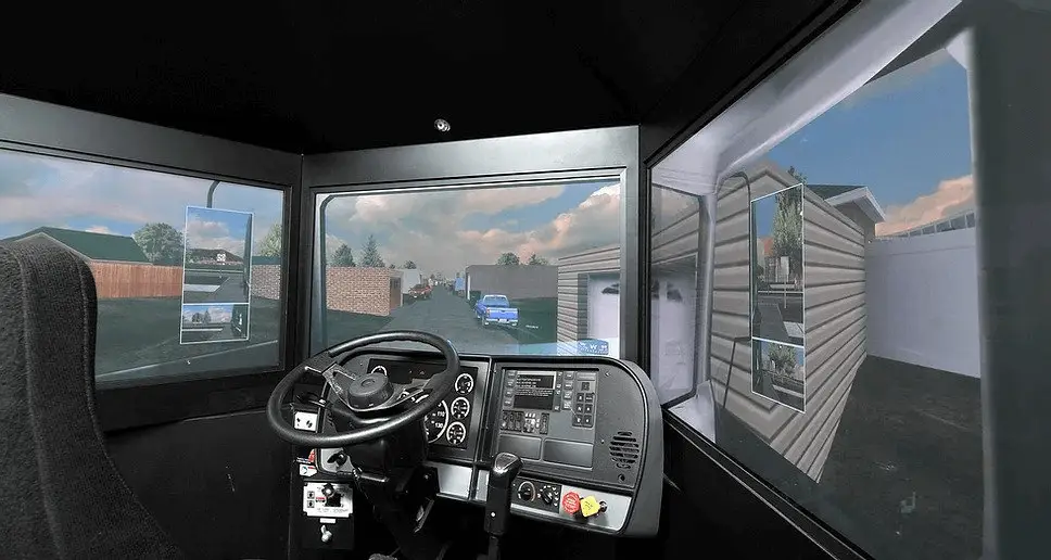 View from inside a driving simulator showing a steering wheel, control panel, and screens displaying a residential neighborhood with houses, fences, and parked cars.