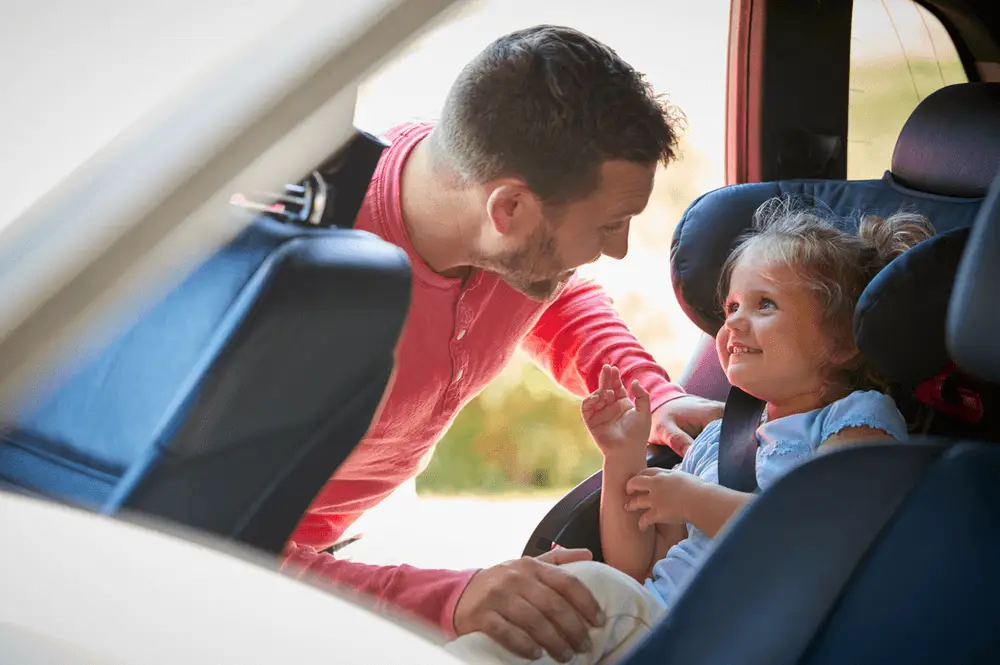 Man-straps-child-into-a-car-seat-DriveWise A man leans into a car, smiling and talking to a young girl who is sitting in a car seat, looking up at him and smiling.