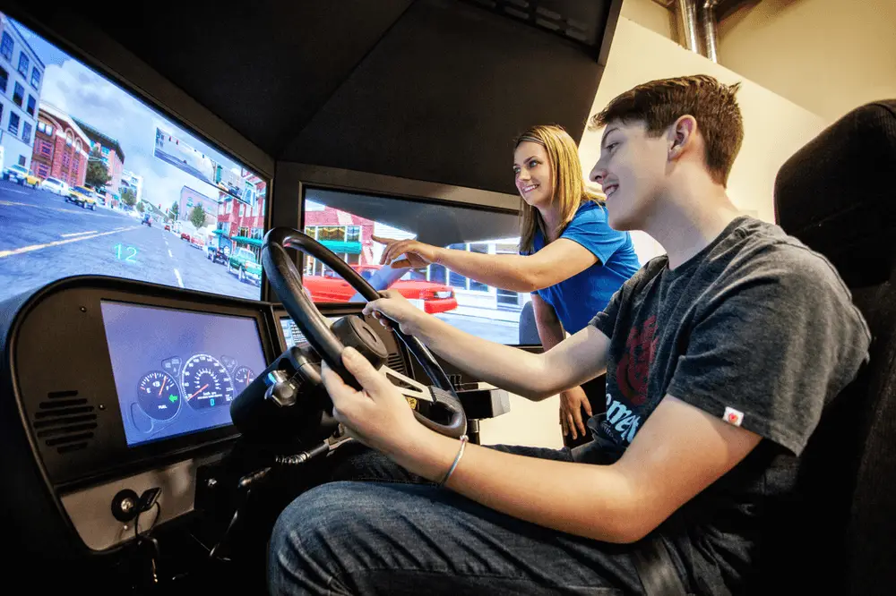 Student-being-instructed-on-a-simulator-DriveWise A young man sits in a driving simulator holding the steering wheel while a woman beside him points at the screen, guiding him. The simulator displays a virtual city street scene.