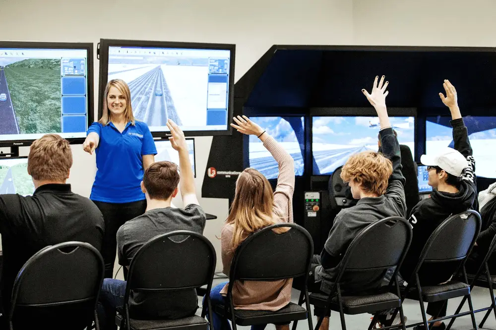 A woman stands in front of computer screens, smiling and pointing at a group of students seated facing her; several students have their hands raised, engaging in an interactive classroom setting.