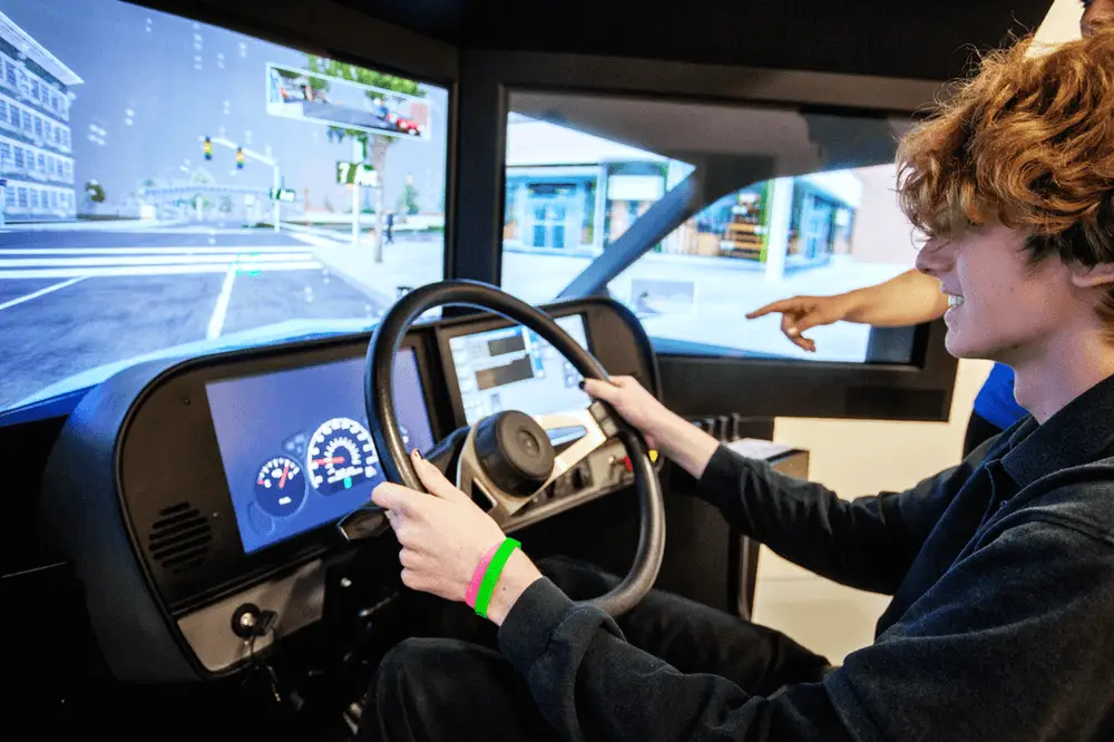 A young person sits at a driving simulator, holding the steering wheel and focusing on virtual road scenes displayed on large screens. Another person’s hand points at something on the simulator dashboard.