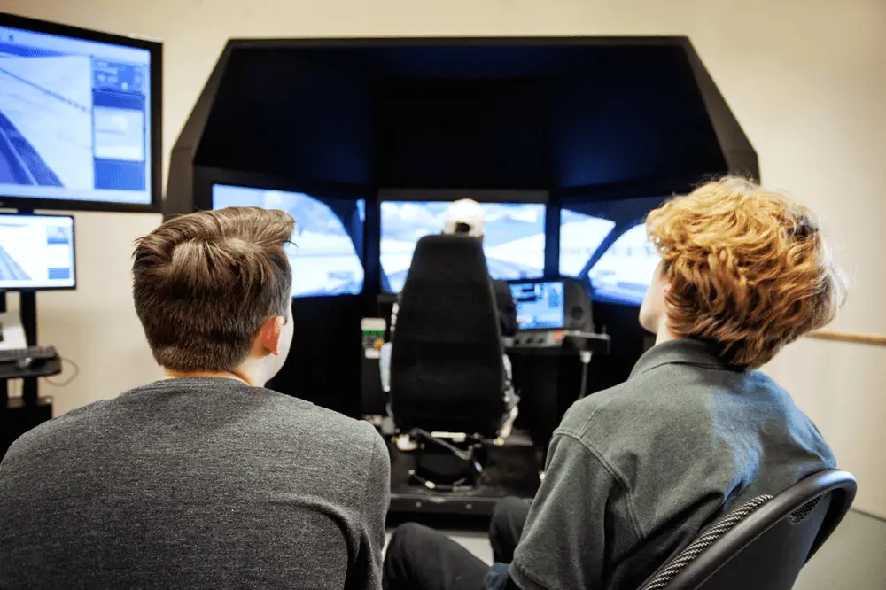 Two young people watch as another person operates a driving simulator with multiple large screens and control panels in a modern, well-lit room. Monitors display simulated road scenes.