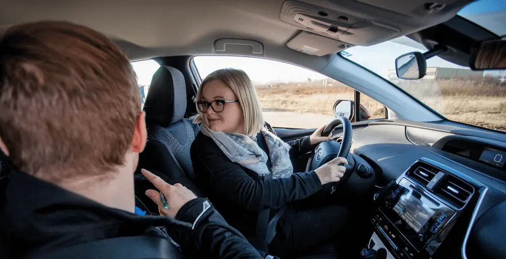 Woman-inside-car-reversing-DriveWise A woman driving a car looks back over her shoulder, possibly reversing, while a man in the passenger seat gestures towards her. The car’s interior and dashboard are visible.