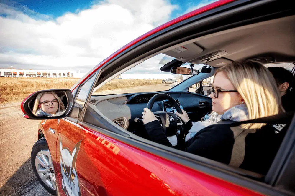 A woman with blonde hair and glasses sits in the driver’s seat of a red car, looking into the side mirror. The car is parked outdoors on a cloudy day, with open fields in the background.