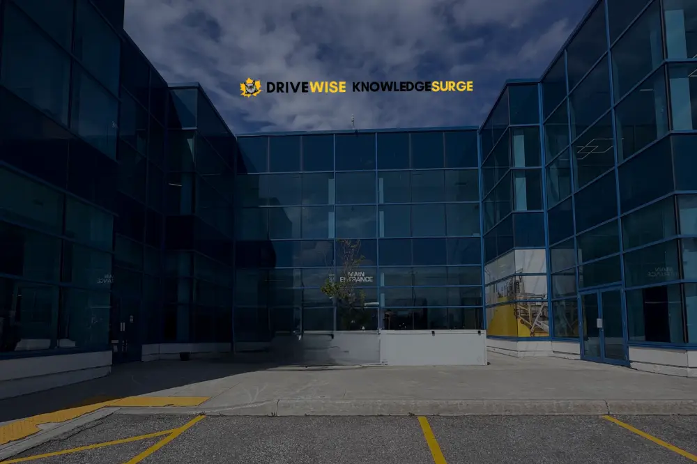 Modern glass building with reflective blue windows under a partly cloudy sky. Above the entrance, a sign reads "DRIVE WISE KNOWLEDGE SURGE" in yellow and black letters. A small tree is planted near the main entrance.