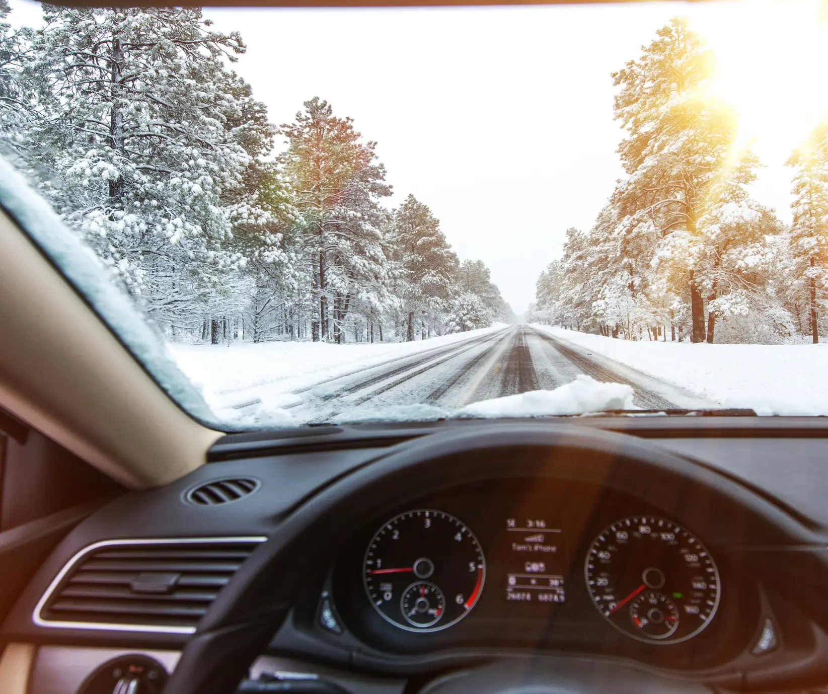View from inside a car driving down a snow-covered road lined with pine trees under a bright winter sun; dashboard and windshield with snow visible in the foreground.