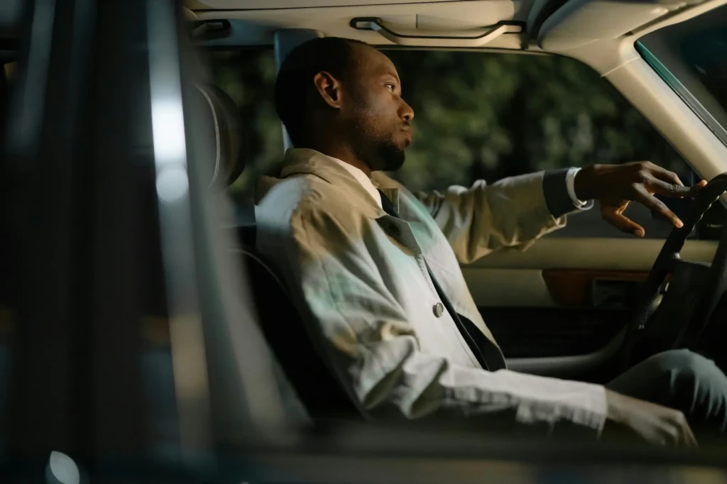 A man wearing a light-colored trench coat sits in the driver’s seat of a car at night, resting his arm on the window and looking thoughtfully out the windshield, as if considering refresher courses to help mature drivers regain confidence.