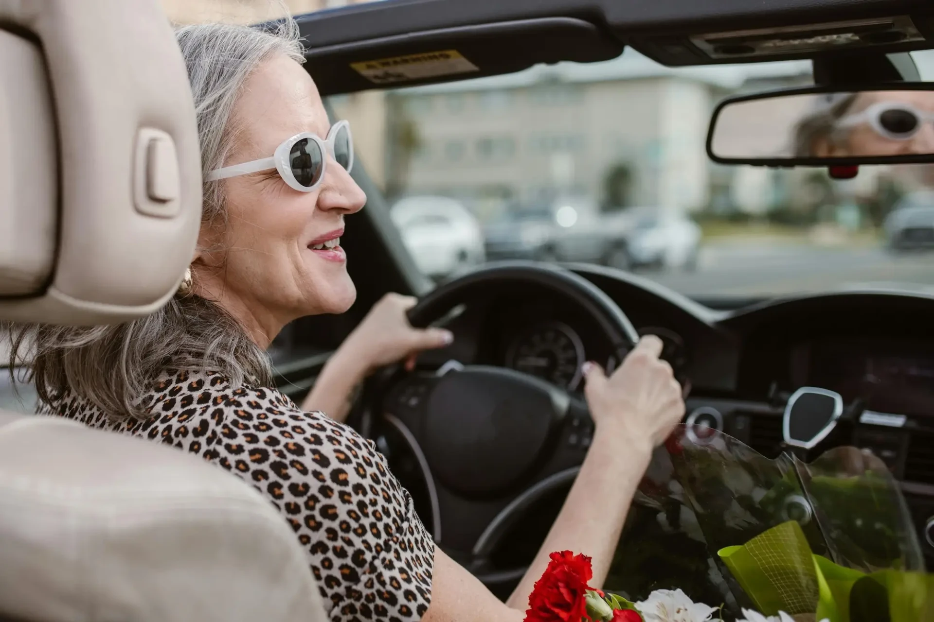 A mature driver with gray hair, wearing white sunglasses and a leopard-print top, smiles as she drives her convertible. A bouquet of flowers rests beside her—proof that mature drivers can regain confidence behind the wheel.