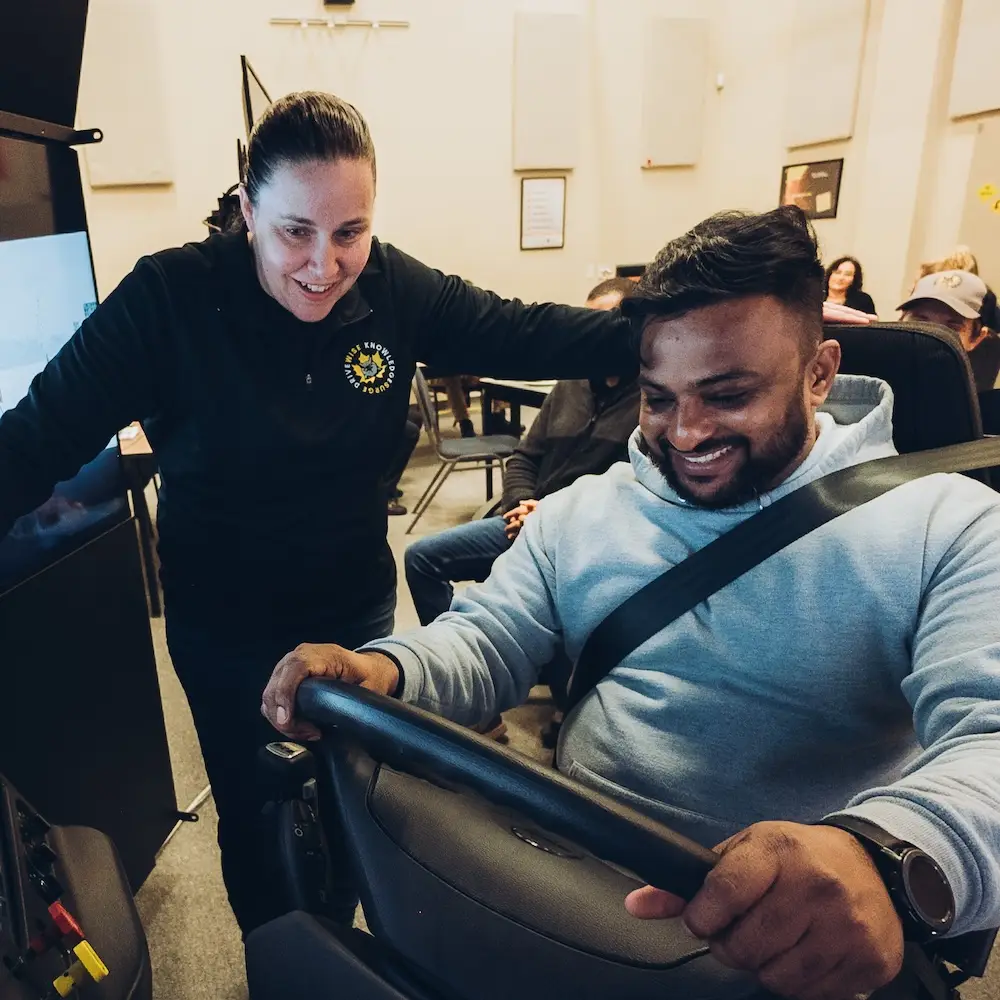 A woman stands beside a smiling man who is sitting at a driving simulator, holding the steering wheel. Other people are seated in the background of the room.