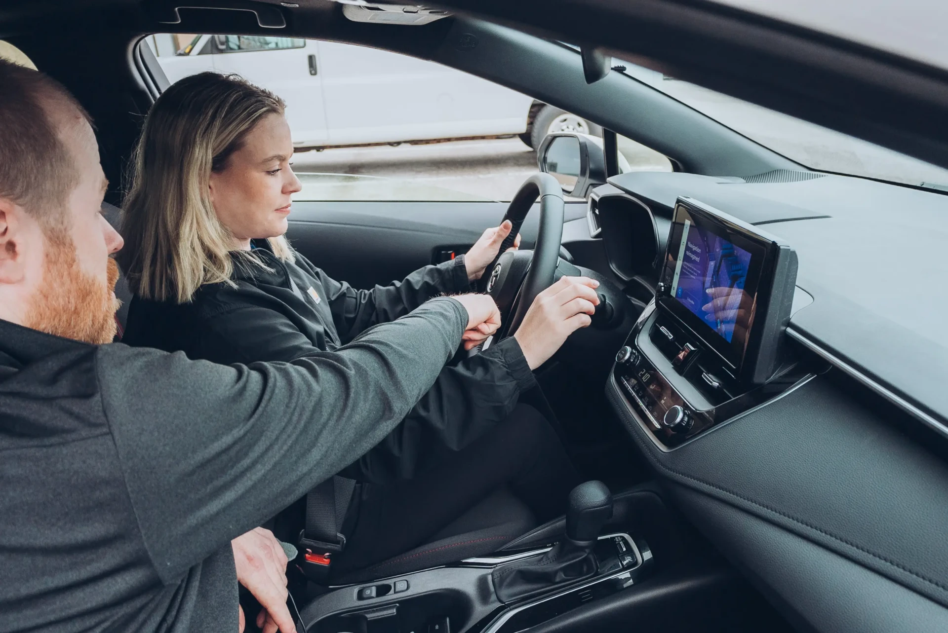 A woman sits in the driver’s seat of a car, turning the ignition key while a man beside her points at the dashboard screen, which displays some information.